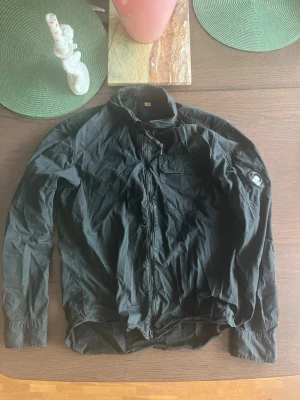 Svart overshirt från CP- company - Snygg svart overshirt från CP- company med dragkedja framtill och klassisk patch på vänster ärm. Overshirten har en bröstficka med lock och knapp samt knappar vid ärmsluten. Tillverkad i ett lätt och slitstarkt material, perfekt för lager-på-lager-stil.