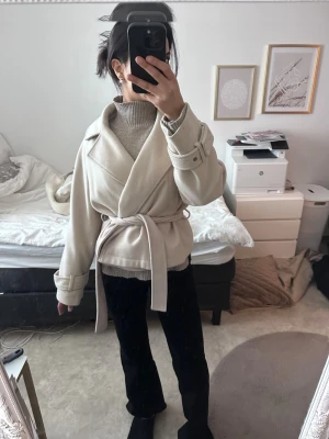 Beige kappa med knytskärp - Stilren beige kappa med bred krage och knytskärp i midjan. Kappan har långa ärmar med dekorativa spännen vid ärmsluten och en avslappnad passform. Perfekt för lager-på-lager och ger en trendig look till hösten och vintern.