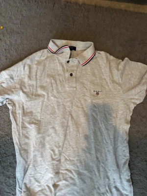 Grå pikétröja från Gant - Klassisk grå pikétröja från Gant med korta ärmar och krage med röd och vit randig detalj. Två knappar framtill och diskret Gant-logga broderad på bröstet. Perfekt till jeans eller shorts.