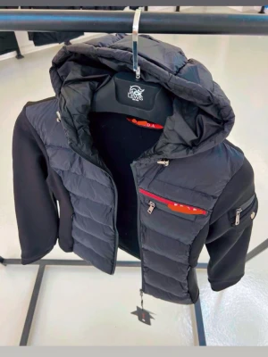 Svart pufferjacka med huva och detaljer - Snygg svart pufferjacka från  med vadderad front och huva. Jackan har dragkedja, röda detaljer på bröstfickan och en extra ficka på ärmen. Ärmarna är i mjukare material för extra komfort. Perfekt 