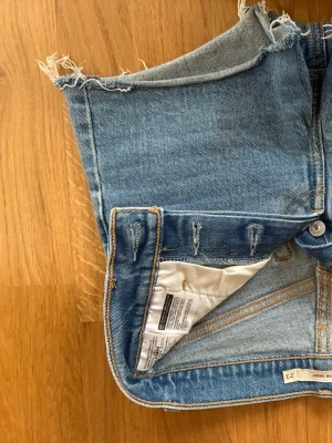 Levi's blå jeansshorts hög midja - Säljer ett par klassiska blå jeansshorts från Levi's med hög midja och råa, fransiga kanter. Shortsen har två bakfickor, knappgylf och den ikoniska läderlappen bak. Perfekta till sommaren!