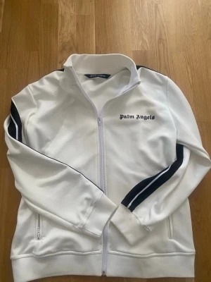 Vit Palm Angels ziptröja med svarta ränder - Snygg vit ziptröja från Palm Angels med svarta ränder längs ärmarna och logga på bröstet. Tröjan har hög krage, dragkedja framtill och fickor med dragkedja. Perfekt för en sportig och trendig look. Står S men skulle säga att den passas M också väldigt bra. Har några små små fläckar men knappt syns och man märker knappt av dom. Kom med ett prisförslag om du är sugen!