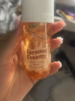 KISS Caramel Creamy Eau de Toilette - Söt parfym från KISS My Body med doft av karamell, amber och träiga toner. Flaskan är genomskinlig med karamellfärgad vätska och har ett vitt lock. Perfekt för dig som gillar gourmanddofter och vill ha något unikt i parfymhyllan.