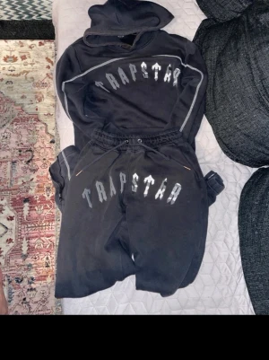 Svart tracksuit  från Trapstar - Svart tracksuit  från Trapstar med stor vit logga över låren. Byxorna har dragsko i midjan och dragkedjor vid fickorna. Sköna och stilrena, perfekta för en avslappnad streetwear-look. Tröjan har några slitande men det går att fixa 