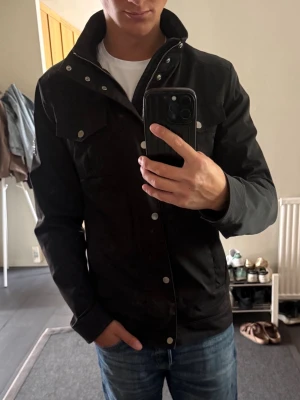 Svart vindjacka från J.Lindeberg - Stilren svart vindjacka från J.Lindeberg med hög krage, dragkedja och tryckknappar framtill. Jackan har flera fickor med lock och en diskret logga på ärmen. Perfekt för dig som vill ha en snygg och funktionell jacka till vardags.