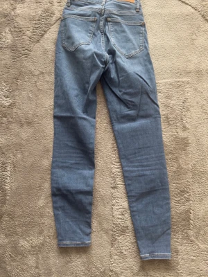 Blå skinny high waist jeans - Säljer ett par blå skinny jeans med hög midja och klassisk femficksdesign. Jeansen har en smal passform hela vägen ner och är tillverkade i stretchigt denimtyg för extra komfort. Perfekta för en trendig och avslappnad stil.
