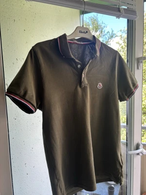 Olivgrön pikétröja från Moncler - Snygg olivgrön pikétröja från Moncler med korta ärmar och klassisk krage. Tröjan har knappar vid halsen och Monclers logga på bröstet. Detaljer i rött och vitt på krage och ärmslut ger en sportig touch. Perfekt till jeans eller shorts.
