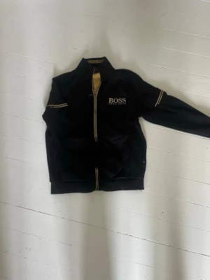 Svart zip-tröja från Hugo Boss - Snygg svart zip-tröja från BOSS med guldig logotyp på bröstet samt guldiga detaljer på ärmarna och vid dragkedjan. Storlek S - fint skick.