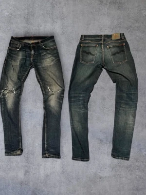 Mörkblå slitna jeans från Nudie - Säljer ett par mörkblå jeans från Nudie med slitningar och hål på ena knät. Jeansen har klassisk femficksdesign, orangea sömmar och en snygg tvättad look. Perfekta för en avslappnad streetstil.