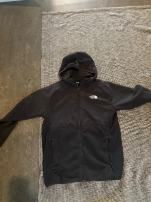Svart hoodie från The North Face - Svart hoodie från The North Face med dragkedja och två fickor framtill. Klassisk logga i vitt på bröstet och mjukt fleecefoder på insidan. Perfekt för en chill och sportig stil. Huva för extra komfort.