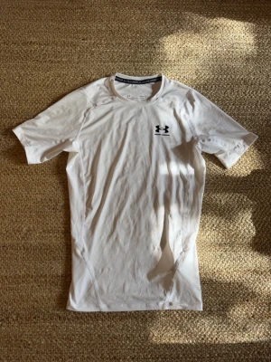 Vit träningströja från Under Armour - Vit kortärmad träningströja från Under Armour med svart logga på bröstet. Tröjan har en tight passform och är tillverkad i ett syntetmaterial som andas (musclefit). Knappt använd. 