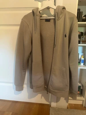 Beige hoodie från Polo Ralph Lauren - Snygg beige hoodie från Polo Ralph Lauren med dragkedja och två fickor framtill. Klassisk logga broderad på bröstet. Mjuk insida och justerbar huva med snören. Perfekt för en avslappnad och stilren look.