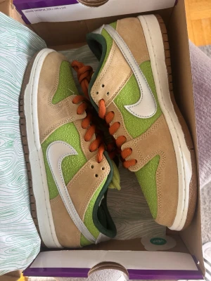 Nike SB Dunk Low Grön/Beige - Nike SB Dunk Low sneakers med grön textilpanel, beige mockadetaljer och vit swoosh. Skorna har orangea skosnören, mörkgrön insida och klassisk gummisula. Perfekt för dig som gillar unika färgkombos och streetwear vibes.