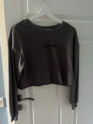 Grå croppad sweatshirt 'c'est la vie' - Säljer en grå croppad sweatshirt med trycket 'c'est la vie' på bröstet. Tröjan har rund halsringning, långa ärmar och rå kant nertill för en avslappnad vibe. Perfekt till jeans eller kjol för en chill look.