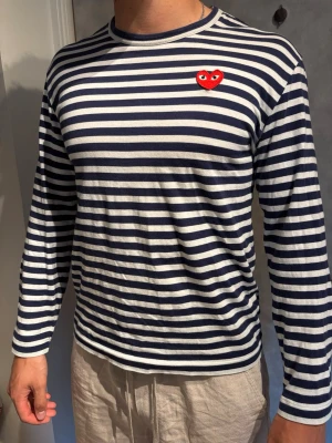 Randig långärmad t-shirt Comme des Garçons - Säljer en långärmad t-shirt från Comme des Garçons med marinblå och vita ränder samt det klassiska röda hjärta-märket på bröstet. Tillverkad i mjuk bomull och har en avslappnad passform. Perfekt för dig som gillar stilrena och ikoniska plagg.