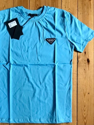 Blå t-shirt från Prada - Snygg blå t-shirt från Prada med klassisk rund hals och korta ärmar. På bröstet sitter en ikonisk svartvit Prada-logga i form av en triangel. Tillverkad i mjuk bomull som känns skön mot huden. Perfekt för dig som gillar stilrena och exklusiva plagg.