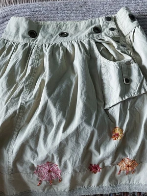 Beige kjol med broderade paraplyer - Säljer en beige kjol med coola metallöljetter i midjan, ficka framtill och broderade paraplyer i rosa, orange och rött längst ner. Kjolen har en avslappnad passform och är perfekt för dig som gillar unika detaljer.