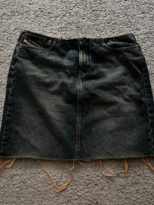 Svart jeanskjol från Diesel - Cool svart jeanskjol från Diesel med råa kanter och klassiska fickor fram. Kjolen har en rak passform och slitna detaljer längst ner för en edgy look. Perfekt att styla med sneakers eller boots för en avslappnad vibe.