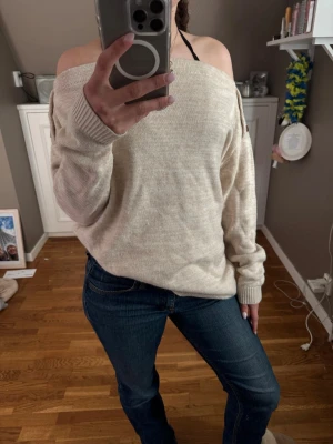 Beige off shoulder stickad tröja - Säljer en beige stickad tröja med off shoulder-modell och dekorativa knappar på ena axeln. Tröjan har långa ärmar och ribbade muddar vid ärmslut och nederkant. Perfekt för en avslappnad och trendig look.