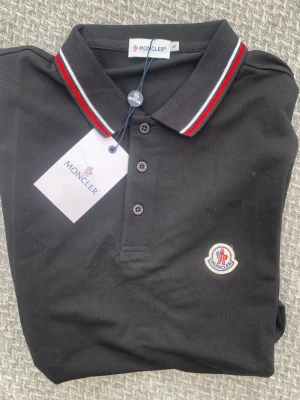 Svart pikétröja från Moncler - Svart pikétröja från Moncler med klassisk krage och knäppning framtill. Kragen har röda och vita ränder som snygg detalj. Tröjan har Moncler-logga broderad på bröstet och är tillverkad i bomull för en bekväm känsla.