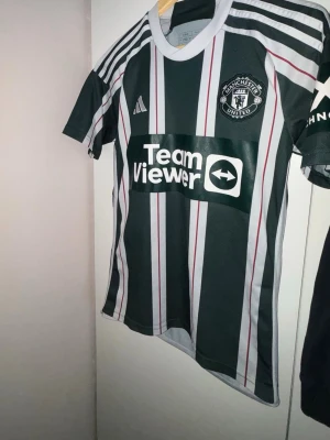 Manchester United matchtröja Adidas - Snygg Manchester United matchtröja från Adidas i svart, vitt och rött med klubbmärke på bröstet och TeamViewer-logga. Tröjan har korta ärmar, rund hals och klassiska ränder. Perfekt för fotbollsträning eller att bära på matchdag.