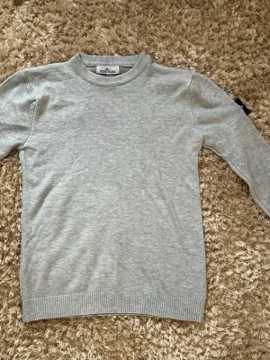 Ljusgrå stickad tröja Stone Island - Snygg ljusgrå stickad tröja från Stone Island med klassisk rund halsringning och ribbade muddar. Tröjan har den ikoniska svarta logotypen med kompass på vänster ärm. Perfekt basplagg för en stilren look.
