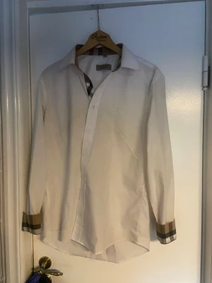 Vit skjorta med rutiga detaljer Burberry - Vit skjorta från Burberry med klassiska rutiga detaljer på insidan av kragen och vid ärmsluten. Skjortan har lång ärm, knappar framtill och är tillverkad i bomull. Perfekt för en stilren och trendig look. m men passar även s jätte bra