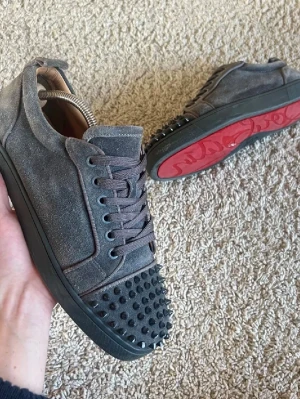 Gråa Louboutin sneakers - Snygga gråa sneakers i mocka från Christian Louboutin med svarta nitar på tån och klassisk röd sula. Skorna har lågt skaft, svarta snören och en cool streetstil. Perfekta för dig som vill sticka ut med en edgy vibe.