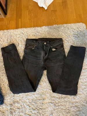 Nudie Jeans - Säljer ett par mörkgråa jeans från Nudie Jeans med klassisk femficksdesign och knappgylf. Jeansen har en smal passform och är tillverkade i stretchigt denimtyg för extra komfort. Perfekta för en clean och stilren look.