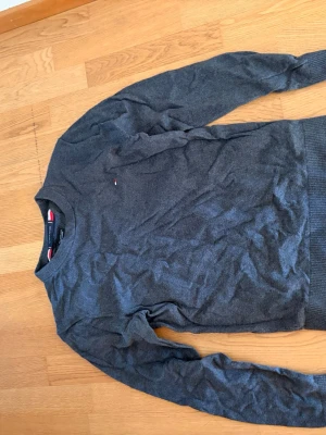 Grå stickad tröja Tommy Hilfiger - Mörkgrå stickad tröja från Tommy Hilfiger i mjuk pima bomullsblandning med cashmere. Klassisk rund halsringning, diskret logga på bröstet och ribbade muddar vid ärmslut och nederkant. Perfekt för en stilren och avslappnad look.