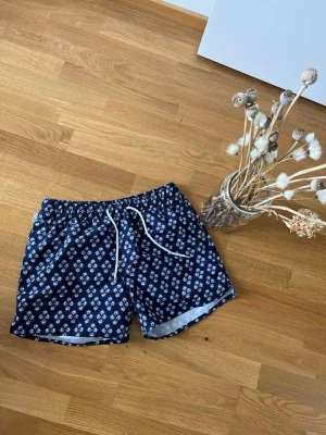 Mönstrade badbyxor från Panos Emporio - Snygga marinblå badbyxor från Panos Emporio med vitt blommönster och vit dragsko i midjan. Klassisk modell med resår i midjan och innerbyxa i mesh. Perfekta för stranden eller poolen.