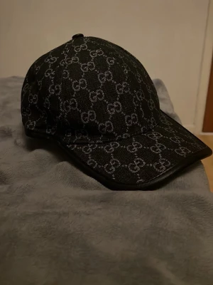 Gucci keps  - Snygg svart keps från Gucci med det klassiska GG-monogrammet i vitt över hela kepsen. Kepsen har böjd skärm och svarta detaljer längs kanten. Tillverkad i ett slitstarkt tyg som ger en exklusiv känsla och passar perfekt till streetwear-looken. Den är helt ny! 