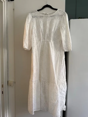 Vit broderad långklänning med snörning - Romantisk vit långklänning från Italien i bomull med broderade spetsdetaljer och vackert hålmönster. Klänningen har trekvartsärm, markerad midja och öppen rygg med snörning. Perfekt för sommarens alla tillfällen.