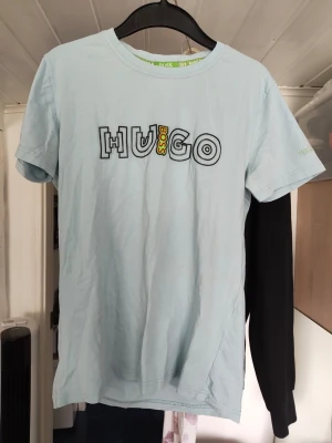 Ljusblå/ turkos Hugo Boss t-shirt - Snygg ljusblå/turkos t-shirt från Hugo Boss med ett stort grafiskt tryck på bröstet där det står 'HUGO BOSS' i svart och gult. Klassisk rund halsringning och korta ärmar. Perfekt för dig som gillar stilrena plagg med en twist.