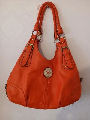 Orange handväska med flätade handtag - Snygg orange handväska från Mulberry med flätade handtag och guldfärgade detaljer. Väskan har en rund form, dekorativa spännen och en metallplatta med Mulberry-loggan på framsidan. Baksidan har ett fack med dragkedja och en matchande liten väska som hänger på kedja.