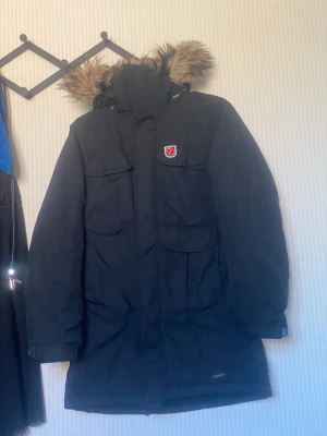 Svart parkas från Fjällräven - Säljer en svart parkas från Fjällräven med avtagbar huva och fuskpäls. Jackan har flera stora fickor framtill, hög krage och klassisk Fjällräven-logga på bröstet. Perfekt för kalla dagar och riktigt snygg streetstil.