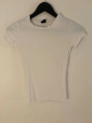 Vit basic t-shirt med rund hals - Enkel och stilren vit t-shirt med rund halsringning och korta ärmar. Modellen är figurnära och passar perfekt till alla typer av outfits. Tillverkad i mjukt bomullsmaterial för en bekväm känsla hela dagen.