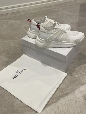 Vita Moncler sneakers med meshdetaljer - Snygga vita sneakers från Moncler med stilrena meshpaneler och detaljer i läder. Skorna har en chunky sula och coola band bak med Monclers signaturränder. Perfekta för dig som gillar exklusiva och trendiga sneakers.