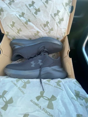 Svarta sneakers från Under Armour - Snygga helsvarta sneakers från Under Armour med diskret logga på sidan. Skorna har en sportig design med mesh-ovandel och tjock sula för extra komfort. Perfekta för dig som gillar en clean och stilren look.