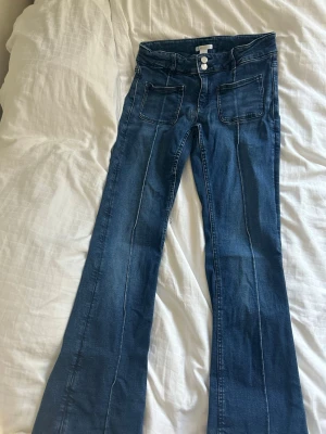 Blå utsvängda jeans med fickor - Snygga blå jeans med utsvängda ben och markerade sömmar. Jeansen har två stora fickor fram, dubbla knappar i midjan och klassiska bakfickor. Perfekta för dig som gillar retrostil och vill ha ett par jeans med coola detaljer. Har en lagning fram vid gylfen. 