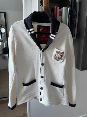 Vit kofta från Morris med emblem - Snygg vit kofta från Morris med mörkblå detaljer, knappar framtill och två fickor. Har ett broderat emblem på bröstet och en klassisk sjalkrage. Perfekt för en preppy stil och passar till både jeans och chinos. Lite större i midjan som man ser på bilderna 