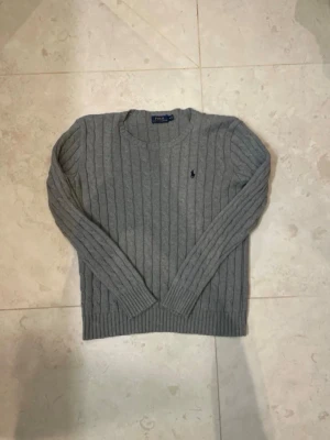 Grå kabelstickad tröja Ralph Lauren - Är mer än Small än en Medium Kabelstickad grå tröja från Ralph Lauren med rund halsringning och klassisk broderad logga på bröstet. Tröjan har långa ärmar och ribbade muddar vid ärmslut och nederkant. Perfekt för en stilren och avslappnad look.