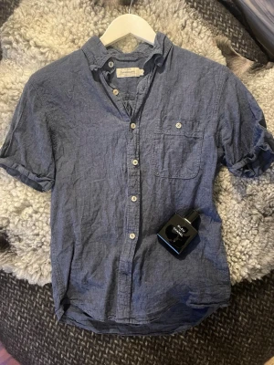 Blå kortärmad skjorta från Denimsmith - Säljer en blå kortärmad skjorta från Denimsmith i mjuk bomull. Skjortan har klassisk krage, bröstficka och vita knappar framtill. Perfekt för en avslappnad och stilren look.