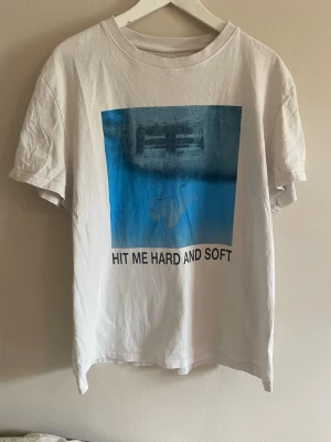 Vit Billie Eilish t-shirt med tryck - Vit t-shirt från Billie Eilish med ett blått grafiskt tryck och texten 'HIT ME HARD AND SOFT' på framsidan. Tillverkad i 100% återvunnen bomull. Den är äkta merch o köpt från hennes hemsida! Använd typ 4 gånger så mycket bra skick💙