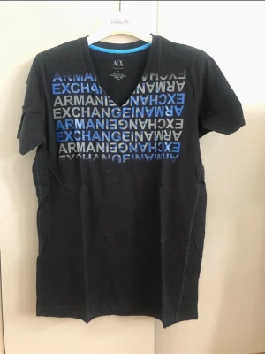 Svart t-shirt från Armani Exchange - Svart t-shirt från Armani Exchange med v-ringning och tryck i blått och grått framtill. T-shirten har korta ärmar och är tillverkad i mjuk bomull. Perfekt för en avslappnad och trendig look.