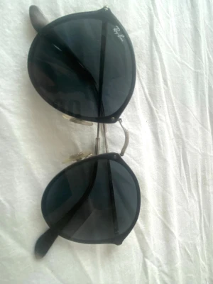 Svarta Ray-Ban solglasögon - Snygga svarta solglasögon från Ray-Ban med klassisk rund form och mörka glas. Metallbågar och näskuddar för extra komfort. Perfekta för att lyfta din stil under soliga dagar.