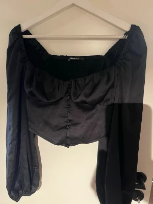 Svart blus med puffärmar från Gina Tricot - Trendig svart blus från Gina Tricot med långa puffärmar och knappar framtill. Toppen har en croppad passform, smockad rygg och en lätt glansig yta. 