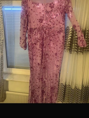 Lång rosa paljettklänning med blommor - Långärmad rosa klänning med blommiga paljettdetaljer och transparenta partier. Klänningen har en elegant, figurnära passform och är täckt av glittrande paljetter som ger ett lyxigt intryck. Perfekt för speciella tillfällen där du vill sticka ut.