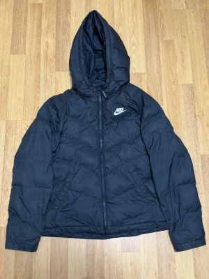 Svart pufferjacka från Nike med huva - Säljer en svart pufferjacka från Nike med dragkedja och stor huva. Jackan har en clean look med Nike-logga på bröstet och är tillverkad i slitstarkt syntetmaterial. Perfekt för kalla vinterdagar och riktigt skön att bära. OBS pytte lite skada se bild. Det är lite lim som har fastnat. Storlek L Barn !