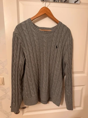 Grå kabelstickad tröja från Polo Ralph Lauren - Kabelstickad grå tröja från Polo Ralph Lauren med rund halsringning och broderad logga på bröstet. Tröjan har långa ärmar och ribbade muddar vid ärmslut och nederkant. Perfekt för lager-på-lager och klassisk stil.